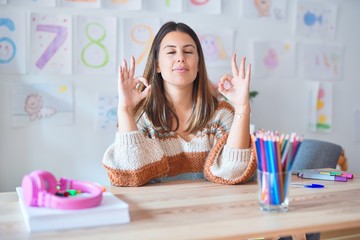 Young beautiful teacher woman wearing sweater and glasses sitting on desk at kindergarten relax and smiling with eyes closed doing meditation gesture with fingers. Yoga concept.