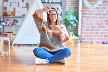 Young beautiful teacher woman wearing sweater and glasses sitting on the floor at kindergarten smiling making frame with hands and fingers with happy face. Creativity and photography concept.