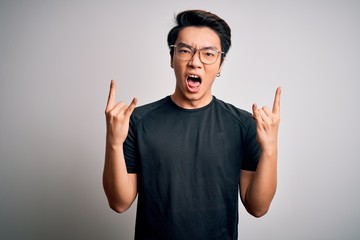 Fototapeta premium Young handsome chinese man wearing black t-shirt and glasses over white background shouting with crazy expression doing rock symbol with hands up. Music star. Heavy concept.