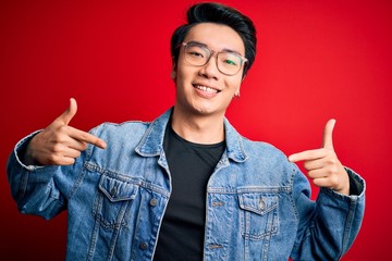 Young handsome chinese man wearing denim jacket and glasses over red background looking confident...