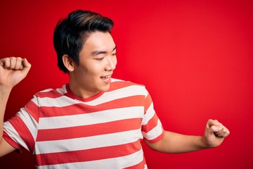 Young handsome chinese man wearing casual striped t-shirt standing over red background Dancing happy and cheerful, smiling moving casual and confident listening to music