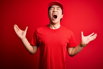 Young handsome chinese delivery man wearing cap standing over isolated red background crazy and mad shouting and yelling with aggressive expression and arms raised. Frustration concept.