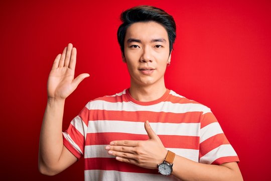 Young handsome chinese man wearing casual striped t-shirt standing over red background Swearing with hand on chest and open palm, making a loyalty promise oath