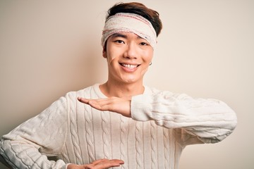 Young handsome chinese man injured for accident wearing bandage and strips on head gesturing with hands showing big and large size sign, measure symbol. Smiling looking at the camera. Measuring