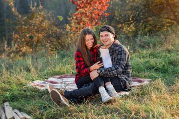 Beautiful girl sits on her boyfriend lap and using smart phone on the lawn in the park. Hipster modern couple have a chatting with their friends sitting in the nature.