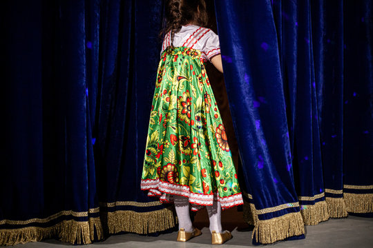 The Child Is Waiting For A Performance On Stage. A Girl Peeks Behind The Curtain. Theatrical Stage With A Blue Velvet Curtain. Costume Embroidered In Folk Patterns. Disturbing Performance. 