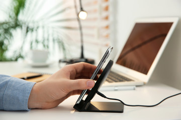 Man putting mobile phone onto wireless charger at white table, closeup. Modern workplace accessory
