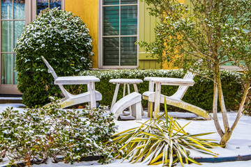 Two white Adirondack chairs with a table between them in the snow in front of a colorful house with green shutters and shrubbery around them