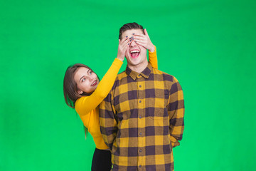 Playful girl covering boyfriend's eyes with hands on green background.