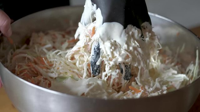 Mixing creamy dressing with crisp shredded cabbage and cut carrots into a delicious homemade coleslaw salad in a chrome bowl wearing a black glove