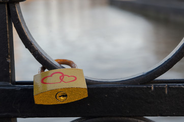 Golden padlock with two red hearts and rusty bracket hangs on black rounded swirls of bridge fence...