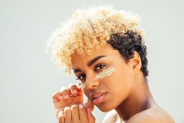 Happy latin brazilian woman with healthy skin applying tone cream on her face on white studio background. she covers bags under the eyes