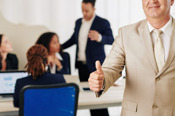Cropped image of middle-aged successful businessman showing thumbs-up, his team discussing project in background