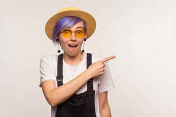 Look there! Portrait of surprised fashionable hipster girl with violet hair in sunglasses and hat pointing to the side, copy space for promotional text, advertisement. white background, studio shot
