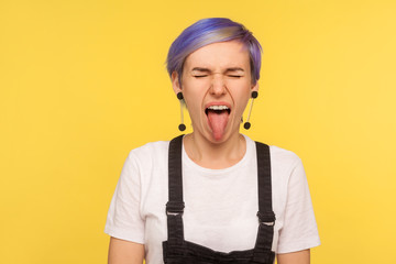 Portrait of naughty hipster woman with violet short hair in denim overalls sticking out tongue and keeping eyes closed, disobedient showing derisive grimace. isolated on yellow background, studio shot