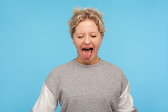 Naughty Woman With Short Hair In Sweatshirt Standing With Closed Eyes And Sticking Out Tongue, Making Derisive Goofy Face, Being Disobedient Unruly. Indoor Studio Shot Isolated On Blue Background