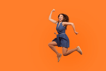 Fototapeta na wymiar Leap to success. Full length of excited energetic motivated brunette woman in denim dress running in air, hurrying to achieve goal, jumping rejoicing victory. studio shot isolated on orange background
