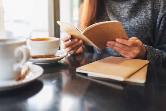Closeup image of a woman holding and reading a book with coffee cup on the table