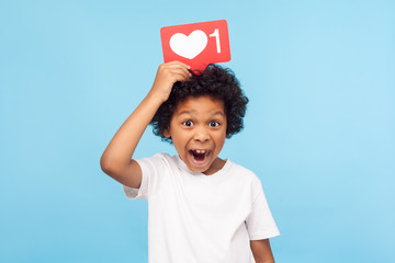 Cute surprised funny little boy with curly hair holding heart Like icon on his head and open mouth in amazement shock, showing button of social network. indoor studio shot isolated on blue background