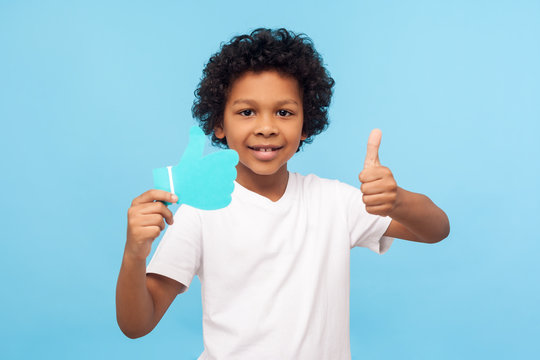Portrait of positive nice preschool boy with curly hair showing thumbs up gesture and holding blue Like icon, appreciates blog comment, trendy children content. indoor studio shot blue background