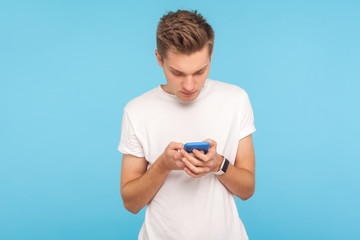 Portrait of young man in white t-shirt using cellphone with focused serious expression, addicted to mobile communication, chatting in social network. indoor studio shot isolated on blue background