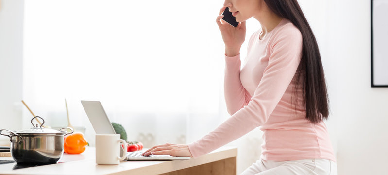 Woman working on laptop and cellphone at kitchen
