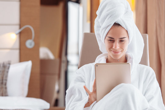 Cheerful young beautiful woman is resting after shower in towel and gown and is looking for training courses on the Internet using a tablet and high-speed Internet. Millennial employment concept - Powered by Adobe