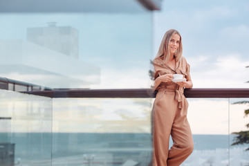 Portrait of a happy smiling caucasian woman in stylish overalls with a cup of coffee meeting the day standing on the hotel terrace with a gorgeous view of the sea and nature