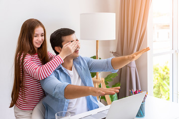 Daughter playing closing her father eye with hand, surprise greeting, while he is working at home with notebook computer