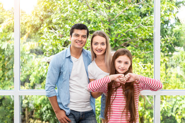 Family, including father mother and daughter, standing and embracing in front of glass window at home, looking at camera