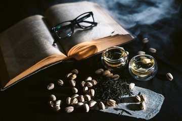 Tea cups at black table with pistachios and old book with glasses at background