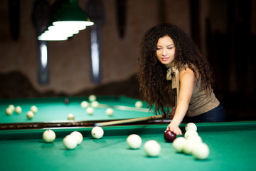 Beautiful girl with voluminous curly hair playing billiards, photo with blurred background
