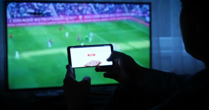 Background of the TV at home (at the bar), the guy orders different snacks, pizza and sweets scrolling through internet stores using mobile phones. Concept: Sport TV.