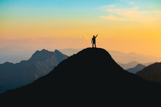 The silhouette of a man with a cheerful raise in his hand on the top of the mountain During sunset time The concept of effort and success