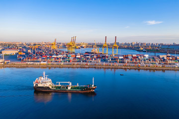 Cargo ship is on the fairway. Sea commercial port. On the pier are containers with goods. Cranes carry out loading and unloading. Transportation of goods by sea. Panorama of the port.
