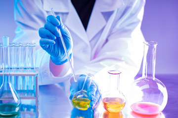 The hands of the chemist in the blue gloves. Laboratory holds a glass rod to stir the solution. On the table are flasks with colorful chemical liquids. Evaluation of experimental results.