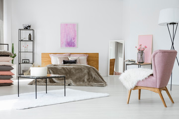 Trendy white bedroom with king size bed, industrial shelf and stylish pink armchair, abstract painting on empty wall