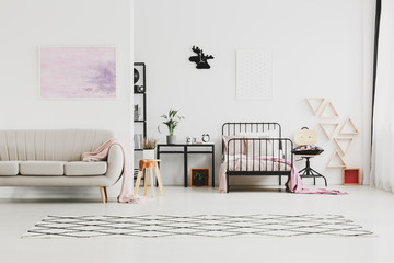 Abstract pastel pink painting above beige stylish couch in kid's bedroom with industrial furniture and patterned rug on the floor