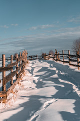 Winter season, landscape picture of a village road covered by snow at sunset, footpath in snow and old timber fence on the side of the road