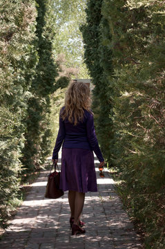 back view of beautiful confident woman with long hair in purple skirt and blouse with bag near green thuja in garden