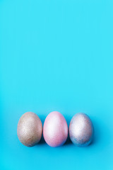 Three colored chicken eggs of pink, silver, golden and blue color on a light blue pastel background. Minimalistic creative classic easter festive vertical flat lay. Copy space for text