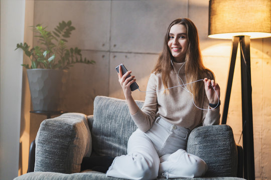 Young female sitting on sofa with smartphone in hand, wearing earphones and listening to music. Girl with mobile phone sending voice messages to friends in social networks, watching stories.