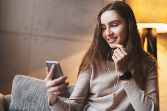Young woman looking at screen of smartphone. Girl with long hair wearing earphones listening to music streaming it on mobile phone. Online app and digital tools with downloaded content.