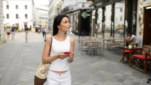 Teen black haired woman in white clothing walking through the calm street, looking around and using smart phone navigation in historical part of Bratislava city. HD VIDEO