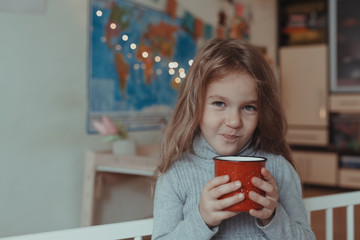 Beautiful little girl drinks cocoa from a red mug on her bed in a cozy room