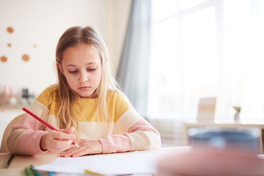 Warm toned portrait of cute little girl drawing pictures or doing homework while sitting at table in home interior, copy space