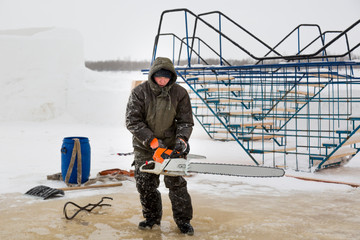 Worker in a green jacket with a hood with a chainsaw in his hands