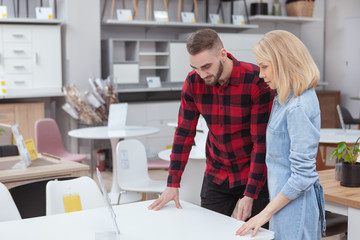 Young couple examining tables on sale at furniture store, copy space. Newlyweds shopping for...