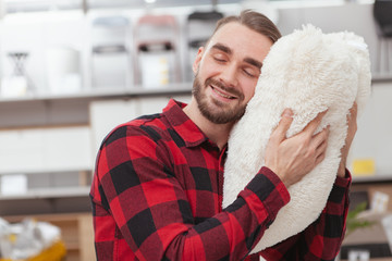Close up of a happy handsome man smiling with his eyes closed, cuddling with fluffy white pillow. Male customer shopping for bedroom goods