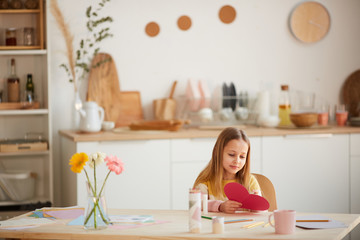 Wide angle portrait of cute girl holding heart-shaped card and smiling while sitting at table in cozy home interior, copy space